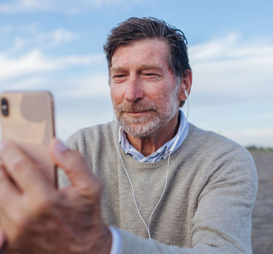 Middle-aged man with brown hair in beige sweater taking a selfie on his phone outside.