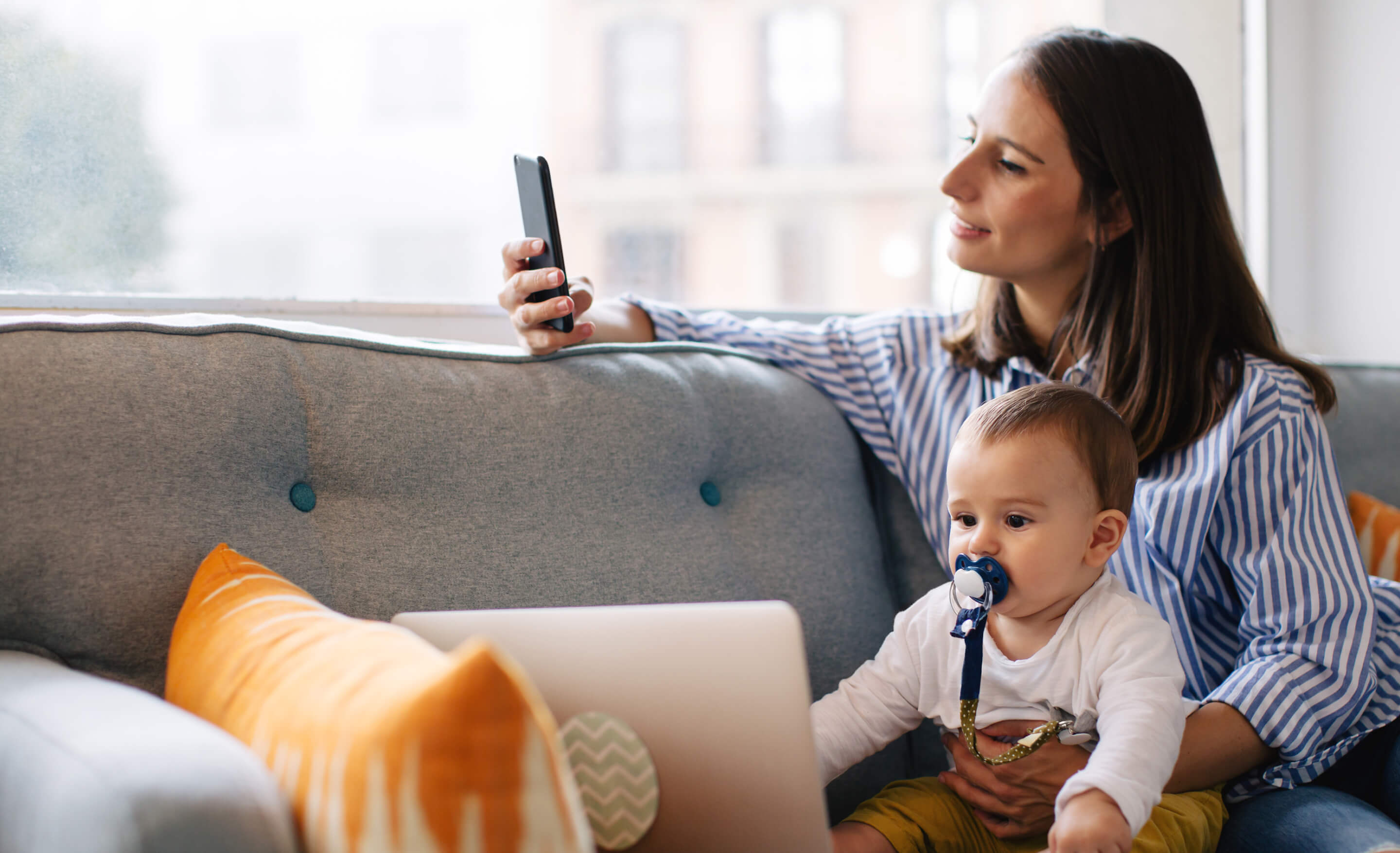 Long-haired brunette woman in stirped blue shirt sits on a couch with a toddler while looking at her phone.