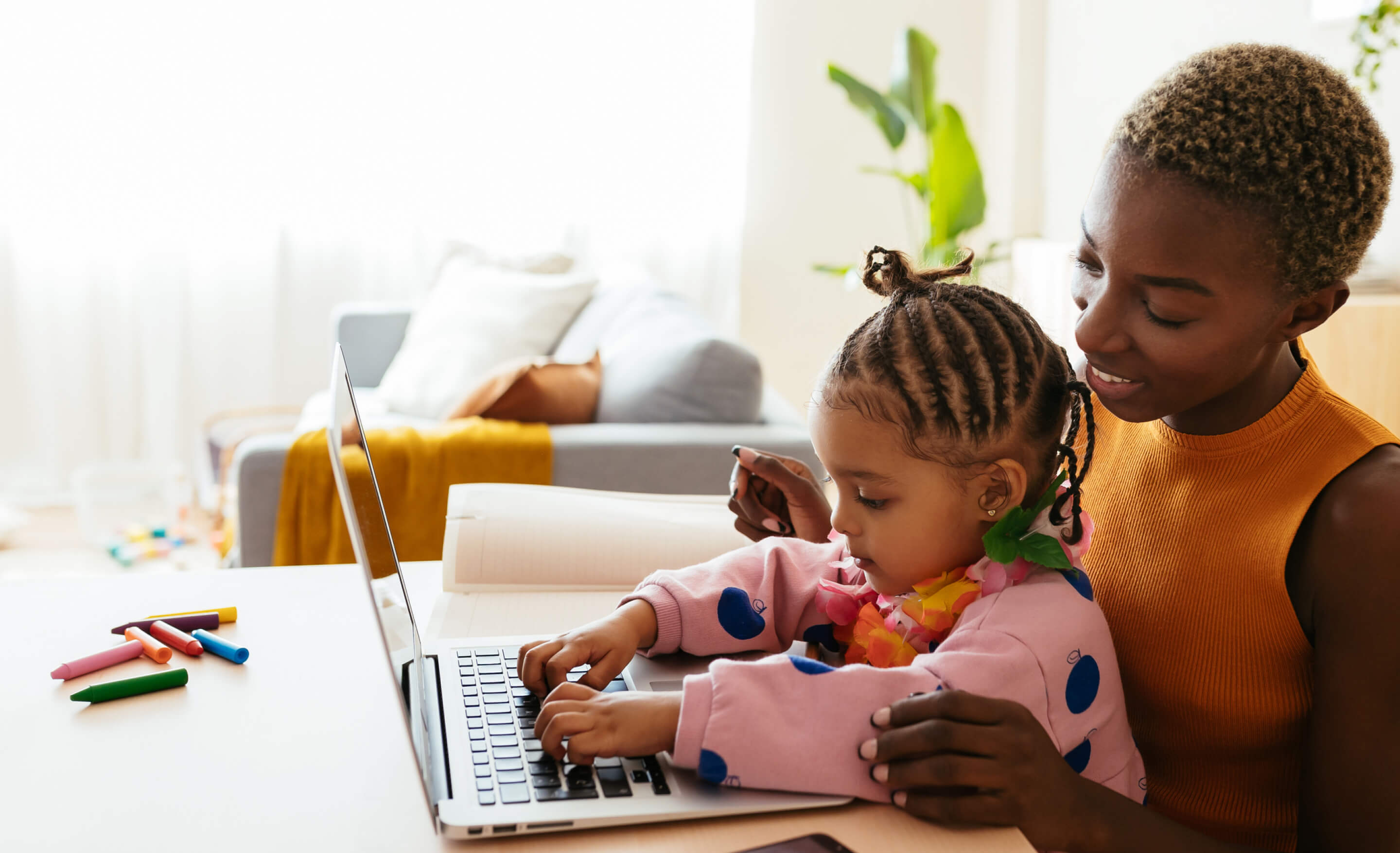 Short-haired black woman smiles as a young girl in her lap types on a laptop.