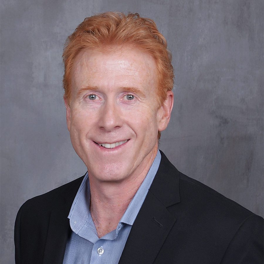 Red-haired man in blue shirt and dark jacket smiling before a gray wall.