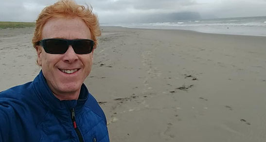 Red-haired man in blue jacket and dark sunglasses smiles outside on the beach.