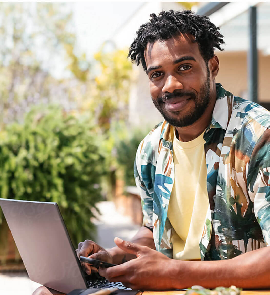 Smiling black man with beard and mustache sits outside with laptop and phone.