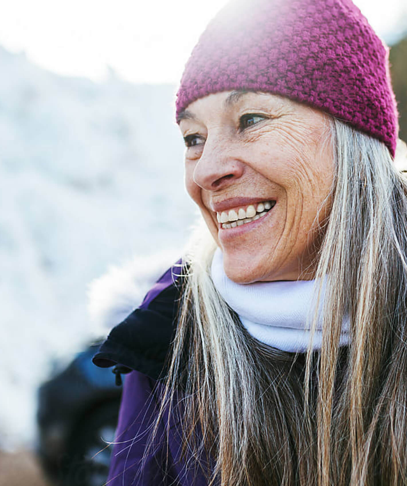 Smiling older woman with gray hair in pink knit cap and purple jacket outside.