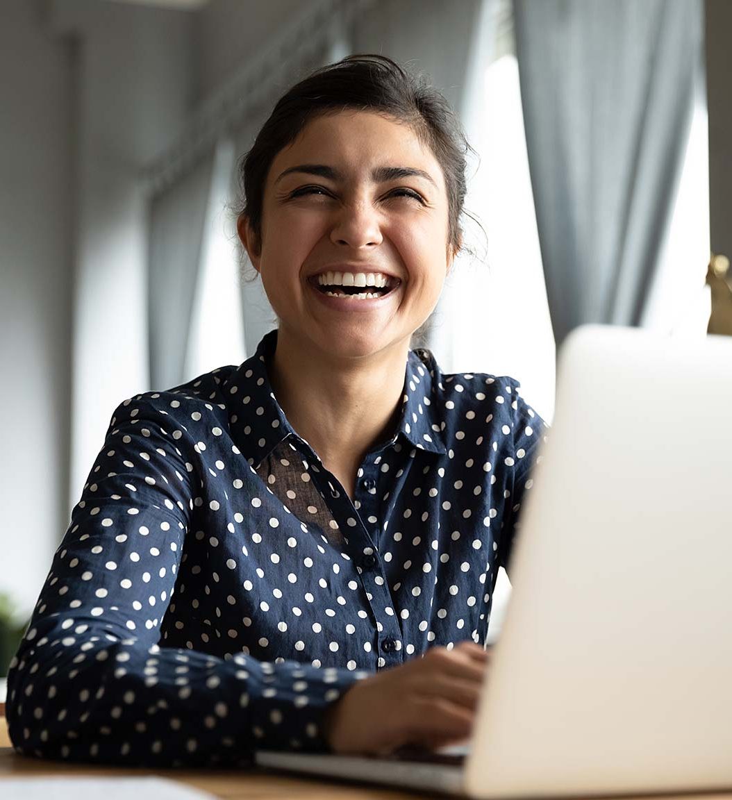 Laughing woman in blue and white polka dot top sits at her laptop in a home.