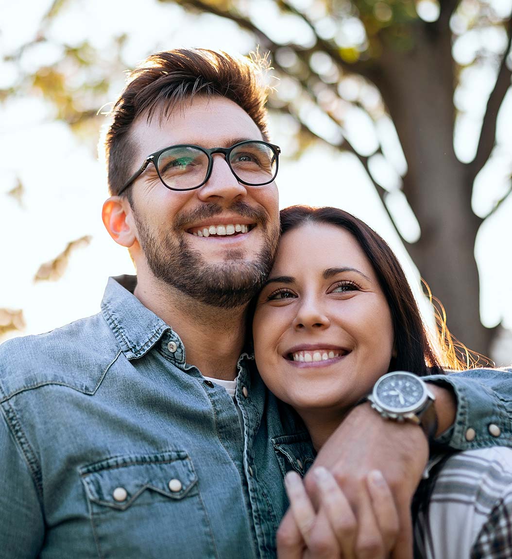 Brunette man with glasses and denim shirt and brunette woman, holding hands and smiling outside by a tree.