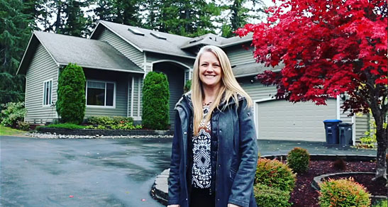 Blonde woman in blue coat smiles in front of a gray house.