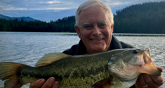 Gray-haired man smiles holding a large fish outside by a lake.