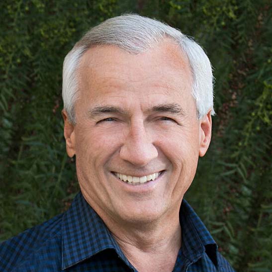 Man with gray hair in blue checkered shirt smiles outside.