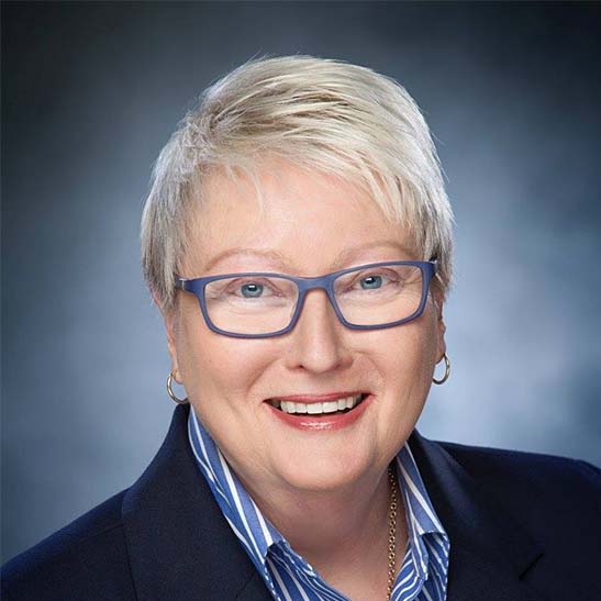 Smiling short-haired blonde woman in blue glasses and blue blazer.